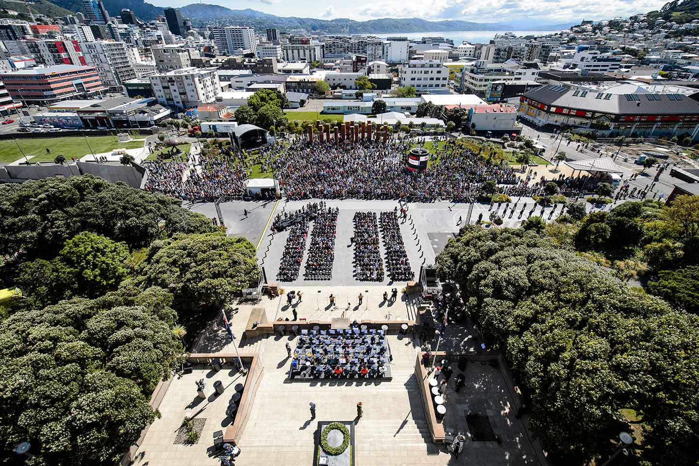 A view from the top of the Carillon tower looking out over Pukeahu park and Wellington on Armistice Day. There is a large group of people on the lawn and on the steps of the Memorial.